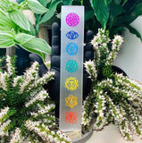 A flat polished chakra bar engraved with seven colorful symbols displayed vertically on a black hand stand.